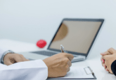 A Dr in a white coat sits at a desk, writing in a notebook. Next to them is an open laptop and a red stress ball. Opposite them, a person in a black jumper sits with their hands folded on the desk. To represent that COTA extends FDA collaboration for RWD cancer research.