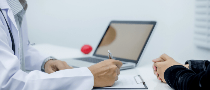 A Dr in a white coat sits at a desk, writing in a notebook. Next to them is an open laptop and a red stress ball. Opposite them, a person in a black jumper sits with their hands folded on the desk. To represent that COTA extends FDA collaboration for RWD cancer research.