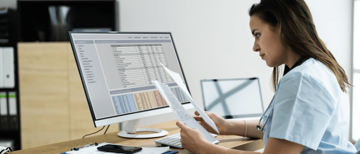 A woman in light blue scrubs wearing a stethoscope around her neck sits at a desk. A computer displays a spreadsheet while the woman holds 2 pieces of paper which she is looking at intently. To represent that Exactis Innovation and PeriPharm partner to enhance RWE studies.