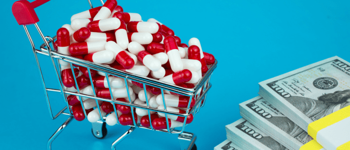 A miniature shopping trolley filled with red and white medicine capsules, next to several stacks of $100 bills. To represent IRA's MDPN Program: a threat to patient access?