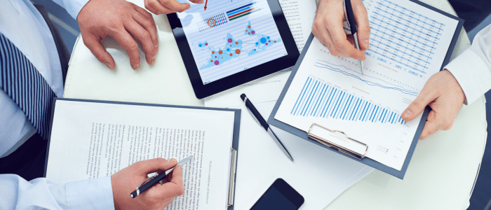 3 people in shirts and one also wearing a tie sit around a table. Two people are writing on documents and the third is looking at graphics on a tablet. To represent that