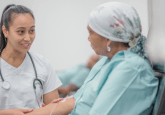 A nurse in light blue scrubs with a stethoscope around her neck holds the arm of a cancer patient receiving treatment. The patient is sitting in a chair and wearing a headscarf patterned with flowers. She has an IV drip in her arm. To represent a partnership to understand RWE of early cancer detection.