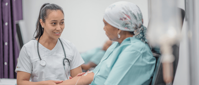 A nurse in light blue scrubs with a stethoscope around her neck holds the arm of a cancer patient receiving treatment. The patient is sitting in a chair and wearing a headscarf patterned with flowers. She has an IV drip in her arm. To represent a partnership to understand RWE of early cancer detection.