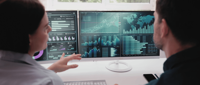 Two people talking in front of 2 computer screens. On the screens are several images of different graphs. To represent that IHI's new playbook enhances data sharing in health research.