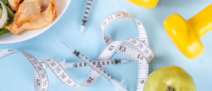 Three needle syringes on a blue background. They are surrounded by measuring tape, two yellow weights, an apple, and a plate with chicken and vegetables.