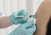 A person wearing blue latex gloves injects a light blue liquid in a syringe into a persons upper arm. They are holding a cotton ball just below the needle. To represent that HHS grants Walgreens $25M for COVID-19 vaccine trial.