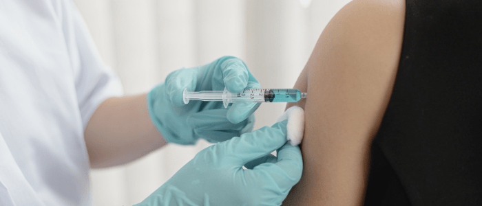 A person wearing blue latex gloves injects a light blue liquid in a syringe into a persons upper arm. They are holding a cotton ball just below the needle. To represent that HHS grants Walgreens $25M for COVID-19 vaccine trial.