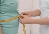 An overweight person in a blue t-shirt has their waist measured by a person in a lab coat, using measuring tape. To represent that a new real-world study unveils trends in GLP-1 RA use.