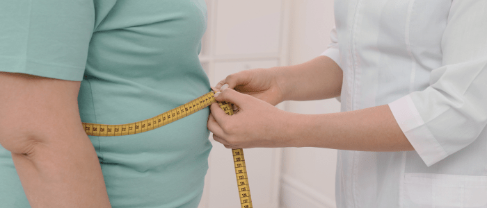 An overweight person in a blue t-shirt has their waist measured by a person in a lab coat, using measuring tape. To represent that a new real-world study unveils trends in GLP-1 RA use.