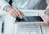 A Dr in a white lab coat sits at a desk, looking at an iPad. On the desk are some files, a sleeve of tablets, two bottles of medication and a fountain pen. To represent that Panalgo and COTA are collaborating to use real-world oncology data to advance cancer care.