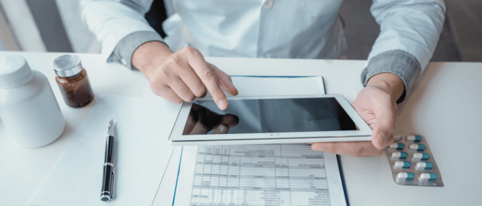 A Dr in a white lab coat sits at a desk, looking at an iPad. On the desk are some files, a sleeve of tablets, two bottles of medication and a fountain pen. To represent that Panalgo and COTA are collaborating to use real-world oncology data to advance cancer care.