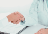 A Dr in a white lab coat wearing a stethoscope around their neck sits at a table with a clipboard on it. They are shaking hands with a person in a white shirt. To represent that Truveta and the CHOICE Institute partner to advance HEOR using RWD.