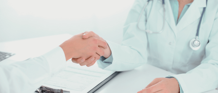 A Dr in a white lab coat wearing a stethoscope around their neck sits at a table with a clipboard on it. They are shaking hands with a person in a white shirt. To represent that Truveta and the CHOICE Institute partner to advance HEOR using RWD.